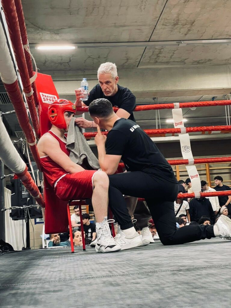 Un boxeur en tenue rouge écoute les conseils de ses coachs dans son coin pendant une pause entre deux rounds, lors d’un combat Boxe amateur à Nivelles.